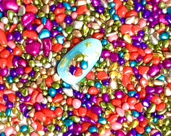 Puede incluir: Primer plano de una uña con base azul claro, detalles dorados y pequeñas cuentas de colores. La uña está rodeada de una mezcla de chispas coloridas en forma de guijarros en tonos rosa, morado, dorado y naranja.