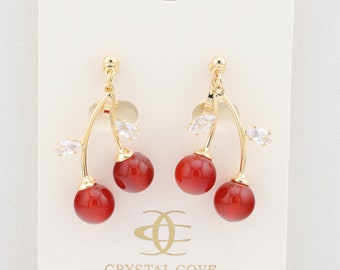 Puede incluir: Pendientes de cereza dorados con detalles de piedras preciosas rojas y hojas de cristal transparente. Los pendientes están en una tarjeta blanca con el texto "Crystal Cove Creations".