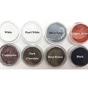 Puede incluir: Ocho peque&ntilde;os recipientes circulares transparentes llenos de pigmentos cosm&eacute;ticos en polvo de colores. Las etiquetas dicen White, Pearl White, Silver Grey, Copper Brown, Cappuccino, Dark Chocolate, Deep Brown y Black, dispuestos en dos filas.