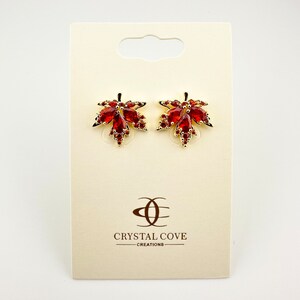 May include: Gold tone earrings with a red crystal maple leaf design. The earrings are on a white card with the text "Crystal Cove Creations".