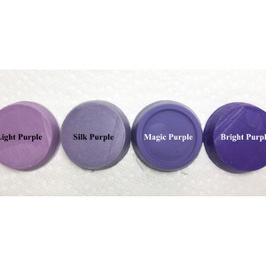 Puede incluir: Cuatro discos circulares de cera o arcilla morada dispuestos en fila. Cada disco tiene una etiqueta con texto en negro o blanco: Light Purple, Silk Purple, Magic Purple y Bright Purple. Los discos muestran diferentes tonos de morado.