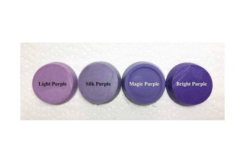 Puede incluir: Cuatro discos circulares de cera o arcilla morada dispuestos en fila. Cada disco tiene una etiqueta con texto en negro o blanco: Light Purple, Silk Purple, Magic Purple y Bright Purple. Los discos muestran diferentes tonos de morado.