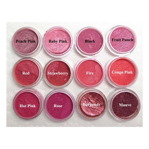 May include: Twelve circular containers of shimmering cosmetic powder arranged in a grid. Each container is labeled with a color name: Peach Pink, Baby Pink, Blush, Fruit Punch, Red, Strawberry, Fire, Congo Pink, Hot Pink, Rose, Burgundy, and Mauve.