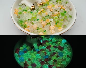 Puede incluir: Un plato de cerámica blanco lleno de pequeñas cuentas translúcidas en forma de rosa en tonos verdes, melocotón y transparente. Las cuentas brillan en verde en la oscuridad. El plato tiene un borde dorado y una figura blanca.