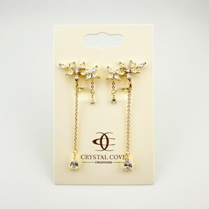 May include: A pair of gold dangle earrings with a floral design and clear crystals. The earrings have a drop-shaped crystal at the end of the chain. The earrings are displayed on a white card with the text "Crystal Cove Creations".