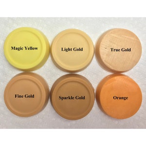 Puede incluir: Seis discos circulares de cera o pigmento de color s&oacute;lido dispuestos en dos filas. La fila superior presenta Magic Yellow, Light Gold y True Gold. La fila inferior muestra Fine Gold, Sparkle Gold y Orange. Cada disco est&aacute; etiquetado con su nombre.