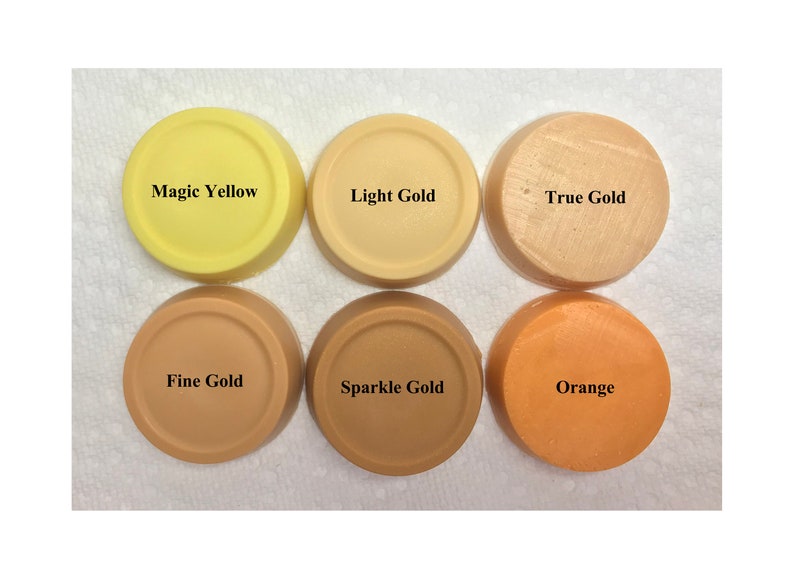 Puede incluir: Seis discos circulares de cera o pigmento de color s&oacute;lido dispuestos en dos filas. La fila superior presenta Magic Yellow, Light Gold y True Gold. La fila inferior muestra Fine Gold, Sparkle Gold y Orange. Cada disco est&aacute; etiquetado con su nombre.