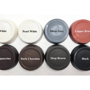 Puede incluir: Ocho botones circulares dispuestos en dos filas, etiquetados con los nombres de los colores: White, Pearl White, Silver Grey, Copper Brown, Cappuccino, Dark Chocolate, Deep Brown y Black. Cada bot&oacute;n tiene un acabado liso y plano.