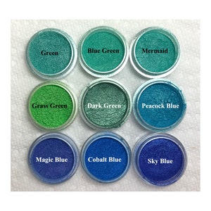 Puede incluir: Nueve recipientes circulares llenos de polvo de mica brillante en varios tonos de verde y azul. Las etiquetas dicen Green, Blue Green, Mermaid, Grass Green, Dark Green, Peacock Blue, Magic Blue, Cobalt Blue y Sky Blue.