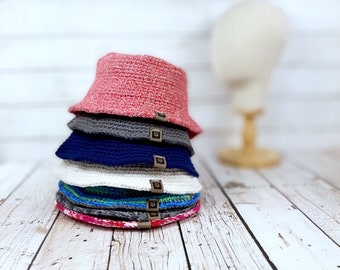 Puede incluir: Una pila de cinco sombreros de cubo de ganchillo en varios colores, incluyendo rosa, gris, azul, blanco y verde. Cada sombrero tiene una pequeña etiqueta marrón con un logotipo.