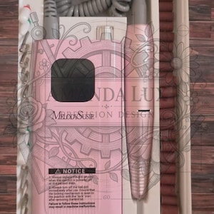 May include: Pink nail drill kit with accessories, including a handpiece, drill bits, and a power supply. The kit is in a white tray and includes the text "AMANDA LU MELODY SUSIE ION DESIGN". A notice is also included.