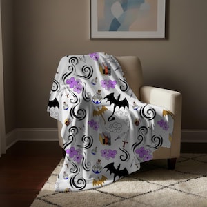 May include: White throw blanket with a fantasy pattern featuring black dragons, purple flowers, potion bottles, books, swords, and swirling black smoke. The blanket is draped over a beige armchair.