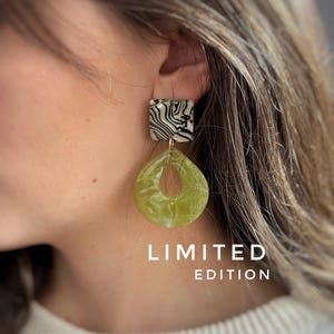 May include: A pair of dangle earrings with a square top and a teardrop-shaped bottom. The square top has a black and white swirled pattern. The teardrop is a translucent green with a hole in the center. The words "LIMITED EDITION" are printed on the image.