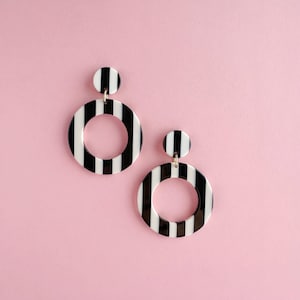 Puede incluir: Dos pares de pendientes a rayas blancas y negras con un diseño circular. Cada pendiente tiene un pequeño perno redondo unido a un aro más grande a rayas. Los pendientes están colocados sobre un fondo rosa claro.