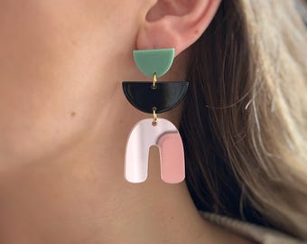May include: Geometric dangle earrings with a three-tiered design. The top tier is a mint green half-circle, followed by a black half-circle, and a pink and silver arch shape at the bottom. The earrings are connected with gold-colored hardware.