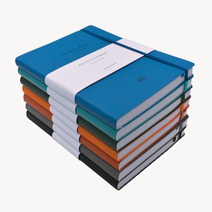 May include: A stack of eight blue, teal, green, orange, and gray hardcover journals with a white band around each one that says "Enlightenment Planner".