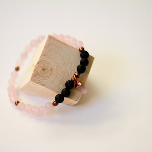 May include: A bracelet made with pink and black beads, with a copper charm. The bracelet is displayed on a wooden block.