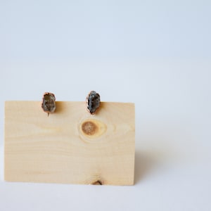 May include: Two raw crystal stud earrings with copper-coloured metal edging are displayed on a light-coloured wooden block. The crystals are a mix of grey and brown tones, set against a white background.