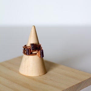 May include: A set of three copper rings with purple gemstone accents displayed on a wooden ring cone.