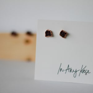 May include: A pair of small, dark brown, irregularly shaped earrings with copper settings. The earrings are displayed on a white card with the text "In Any Kase" written in black ink.