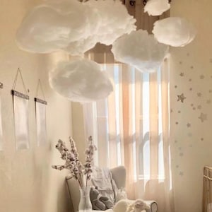 Op de afbeelding: Een kinderkamer in neutrale tinten met een gouden ledikant, een witte fauteuil en een poef. Wolkvormige decoraties hangen aan het plafond. Een kleine bijzettafel bevat een vaas met bloemen en een knuffel.