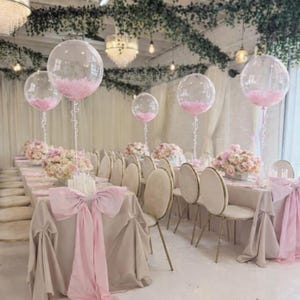 May include: A long banquet table set for an event, featuring beige tablecloths, light pink chair sashes, and floral centerpieces. Clear balloons filled with pink feathers are elevated on thin stems above the table, creating a cohesive decor.