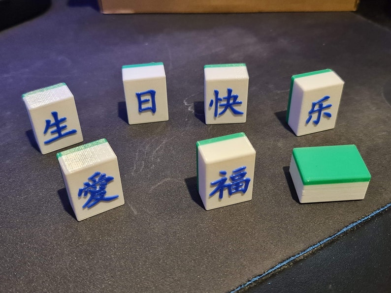 May include: Seven white tiles with green and blue accents. Each tile has a Chinese character written in blue on the top surface. The characters are: 生, 愛, 日, 快, 符, 福, and a blank tile with a green top.