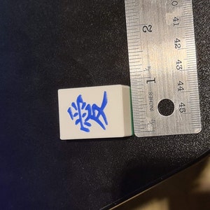 May include: A white tile with blue Chinese characters that read "愛" (love) on it. The tile is about 1 inch wide and 1 inch tall.
