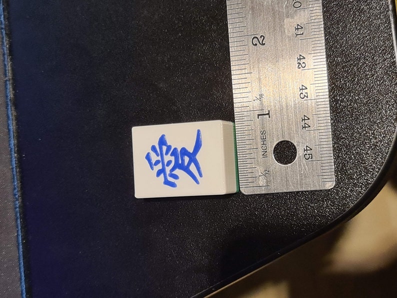 May include: A white tile with blue Chinese characters that read "愛" (love) on it. The tile is about 1 inch wide and 1 inch tall.