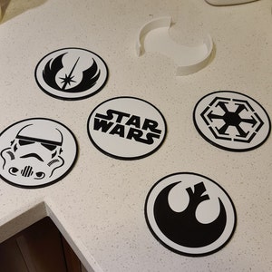May include: Six round white coasters with black Star Wars themed designs. The designs include the Star Wars logo, the Rebel Alliance symbol, the Imperial symbol, a Stormtrooper helmet, and the Jedi Order symbol.