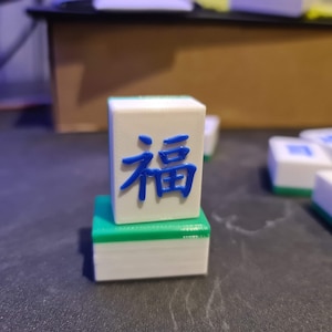 May include: A white and green mahjong tile with the Chinese character "福" (f&uacute;) meaning "fortune" or "good luck" in blue.
