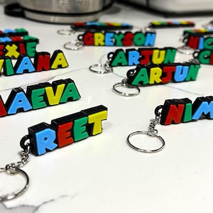 May include: A collection of colourful, personalised keychains with names spelled out in individual letters. The keychains are made of black plastic with colourful letters. The names include Alexie, Aviana, Maeva, Reet, Greyson, Arjun, Nico, Celeste, Nimrit.