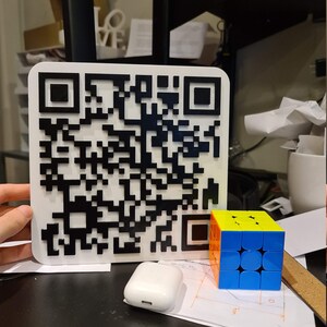 May include: A white square with a black QR code printed on it. A white wireless charging case is next to a blue, yellow, and orange 3x3 Rubik's cube.