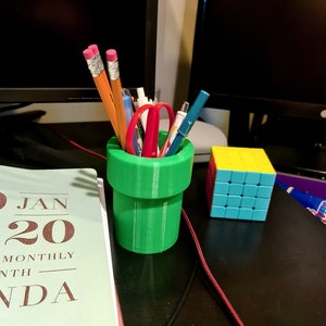 Peut inclure: Un porte-crayons vert imprimé en 3D en forme de tuyau, rempli de crayons, de stylos et d'une paire de ciseaux rouges. Un cube de Rubik bleu et jaune se trouve sur un bureau noir à côté du porte-crayons.