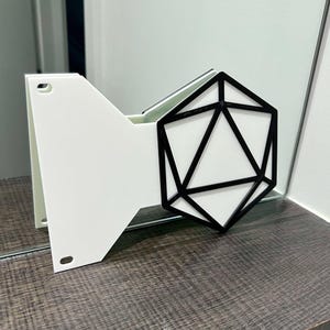 May include: A white and black geometric wall-mounted shelf. The shelf has a white base with a black geometric design on the front. The shelf is mounted on a wooden surface.