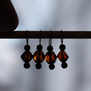 Puede incluir: Cinco cuentas de vidrio ámbar con anillos de metal negro, colgando de una varilla de madera. Las cuentas tienen forma de diamante.