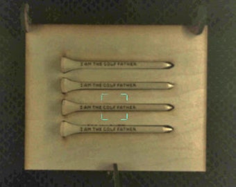 May include: Four wooden golf tees with the text "I AM THE GOLF FATHER" printed on each. The tees are arranged in a row, with the text facing the viewer.