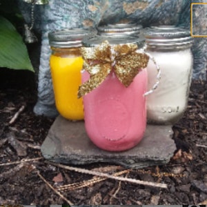 Könnte beinhalten: Drei Glasbehälter mit Deckeln, gefüllt mit farbigem Wachs, stehen auf einem grauen Stein. Die Behälter sind mit einer goldenen Schleife und einem Band verziert. Das Wachs ist gelb, rosa und weiß.