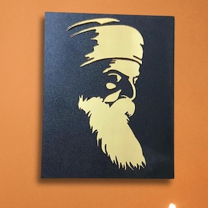 May include: A black wall art piece featuring a gold silhouette of a man with a long beard and turban. Two lit gold candle holders with lotus flower designs sit on a wooden table. A pink flower arrangement is in the background.