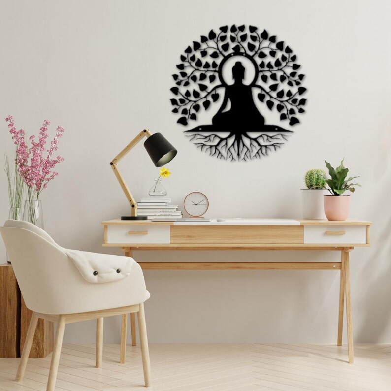 Puede incluir: Arte mural de metal negro que representa a un Buda sentado bajo un &aacute;rbol con ra&iacute;ces. La obra de arte circular est&aacute; montada en una pared blanca. La imagen muestra un escritorio con una l&aacute;mpara, libros y un reloj.