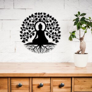 Puede incluir: Arte mural de metal negro que representa una figura de Buda sentado bajo un &aacute;rbol con hojas y ra&iacute;ces. La obra de arte est&aacute; montada en una pared de ladrillo blanco, con una mesa de madera y una planta en maceta en primer plano.