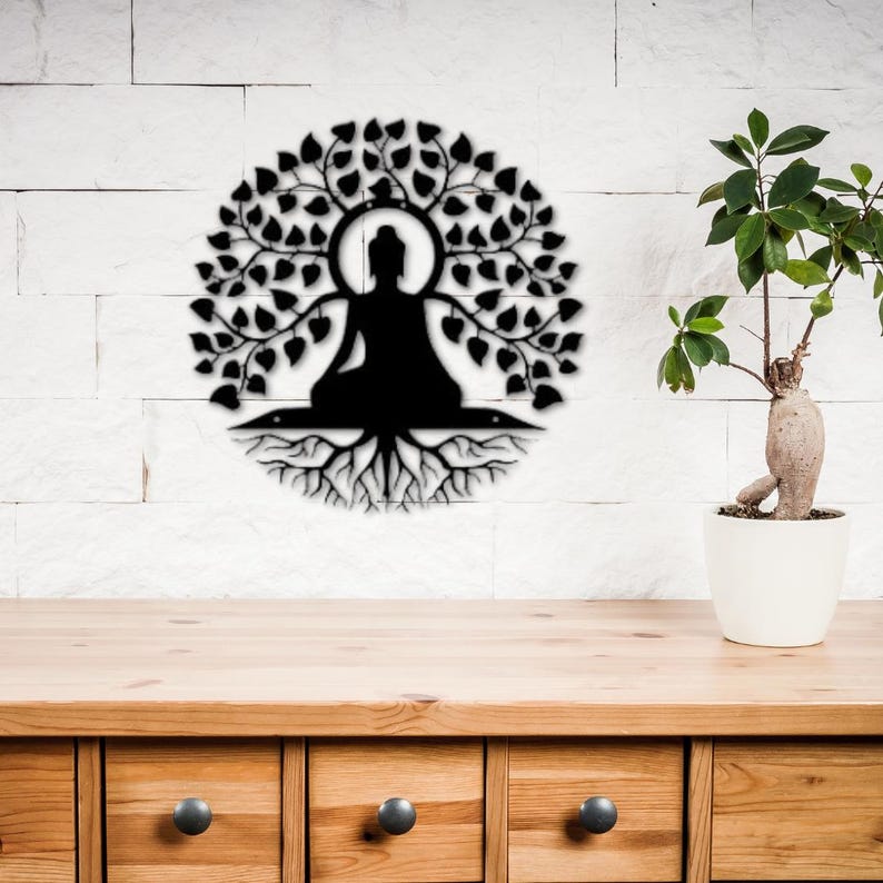 Puede incluir: Arte mural de metal negro que representa una figura de Buda sentado bajo un &aacute;rbol con hojas y ra&iacute;ces. La obra de arte est&aacute; montada en una pared de ladrillo blanco, con una mesa de madera y una planta en maceta en primer plano.