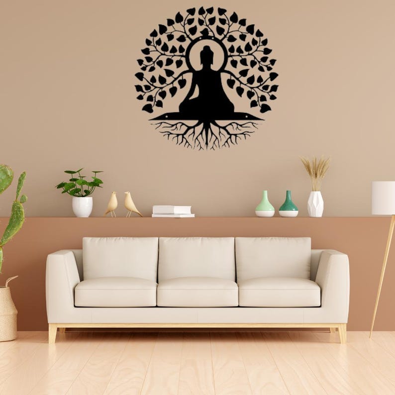 Puede incluir: Una obra de arte mural de metal negro que representa una figura de Buda sentado bajo un &aacute;rbol con un dosel circular de hojas. La obra de arte est&aacute; montada en una pared beige sobre un sof&aacute; blanco, creando un punto focal decorativo.