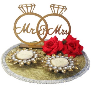 Può includere: Un portafedi dorato con due anelli che recitano "Mr. & Mrs." e due rose rosse su una base circolare dorata e bianca.