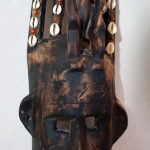 Puede incluir: Una máscara de madera marrón oscuro con un rostro humano tallado y una figura tallada más pequeña en la parte superior. La máscara está decorada con cuentas blancas y naranjas.