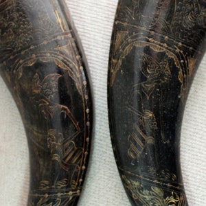 Puede incluir: Dos objetos oscuros en forma de cuerno con intrincados diseños grabados en color dorado. Los cuernos tienen una forma curva y extremos puntiagudos. Los diseños grabados parecen representar figuras y motivos florales. Los cuernos se muestran sobre un fondo claro.