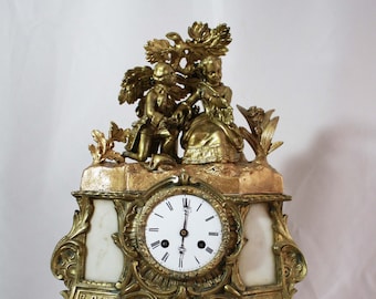 Könnte beinhalten: Eine vergoldete Kaminuhr mit einem weißen Zifferblatt und römischen Ziffern. Die Uhr hat einen dekorativen Aufsatz mit zwei Figuren und einem floralen Design. Der Sockel der Uhr ist mit einem floralen Muster verziert.