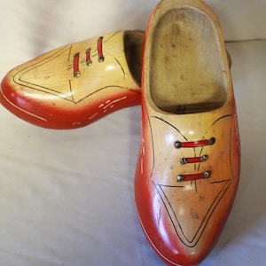 May include: A pair of wooden clogs with a two-tone design. The shoes are painted with a gradient of orange to yellow, with black outlines and red accents for the laces. The clogs have a classic, rounded shape.