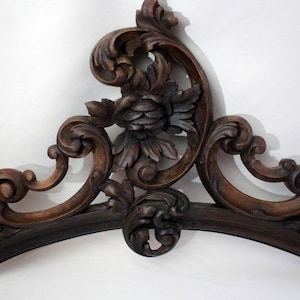 Può includere: Un pezzo decorativo in legno intagliato di colore marrone scuro con un motivo floreale. Il pezzo ha la forma di un arco e presenta dettagli intricati.