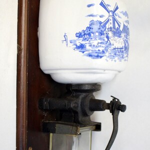 以下が含まれることがあります： 壁掛け式のアンティークコーヒーグラインダー。白いセラミック容器には、青い風車のシーンが描かれています。グラインダーは黒い金属製のメカニズム、ガラス製のキャッチ、木製のハンドルを備えています。濃い色の木材の裏板に取り付けられています。